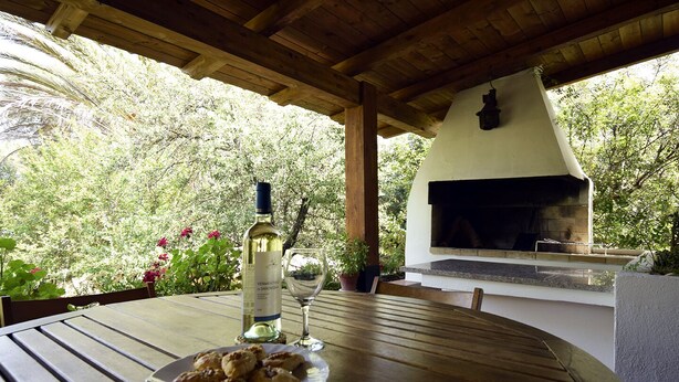 Un espace extérieur aménagé pour les repas, avec une table en bois, une bouteille de vin et un verre. On aperçoit une cheminée en pierre au fond.