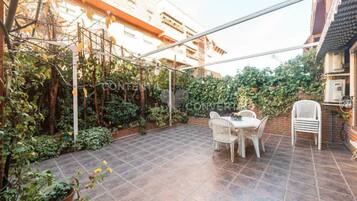 Un patio con suelo de baldosas, sillas blancas y una mesa. Hay plantas y un enrejado con plantas trepadoras.