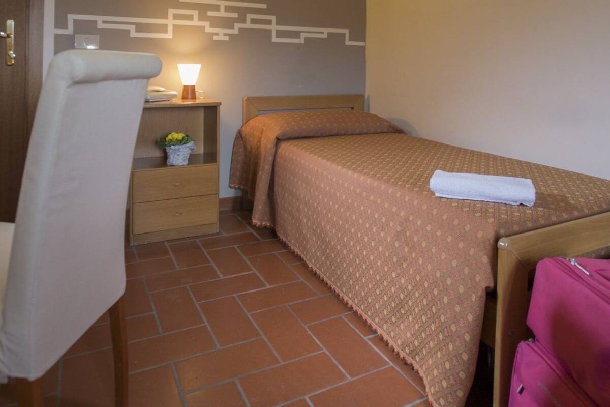 Un dormitorio pequeño y acogedor con una cama individual, una mesita de noche con lámpara, una silla y una maleta.