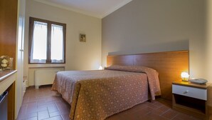 Un dormitori amb un llit, una tauleta de nit, una finestra amb cortines, un radiador i un quadre petit a la paret.