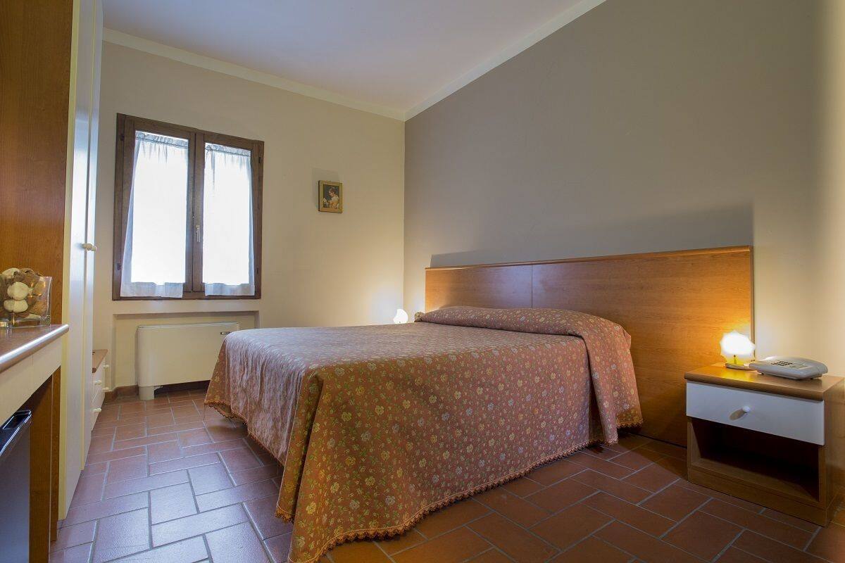 Un dormitorio con cama, mesita de noche, ventana con cortinas, radiador y un pequeño cuadro en la pared.