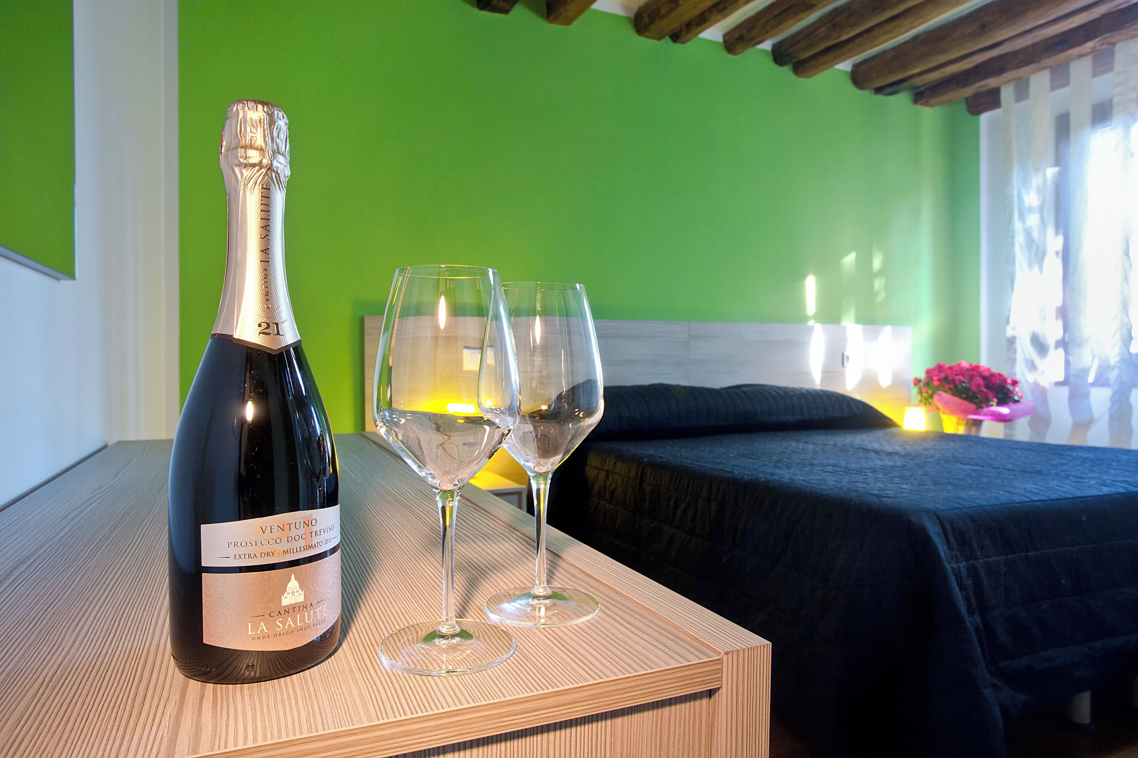 A bottle of wine and two glasses on a table in a room with green walls and a bed with a dark cover.