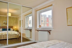 Un dormitorio con un espejo grande, una cama, un radiador y una ventana con vistas a edificios.