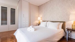 A neatly made bed with white linens and rolled towels, a bedside table with a lamp, and a wardrobe with glass doors. - Charming Flat with A/C & Terrace in Príncipe Real (Lisboa)