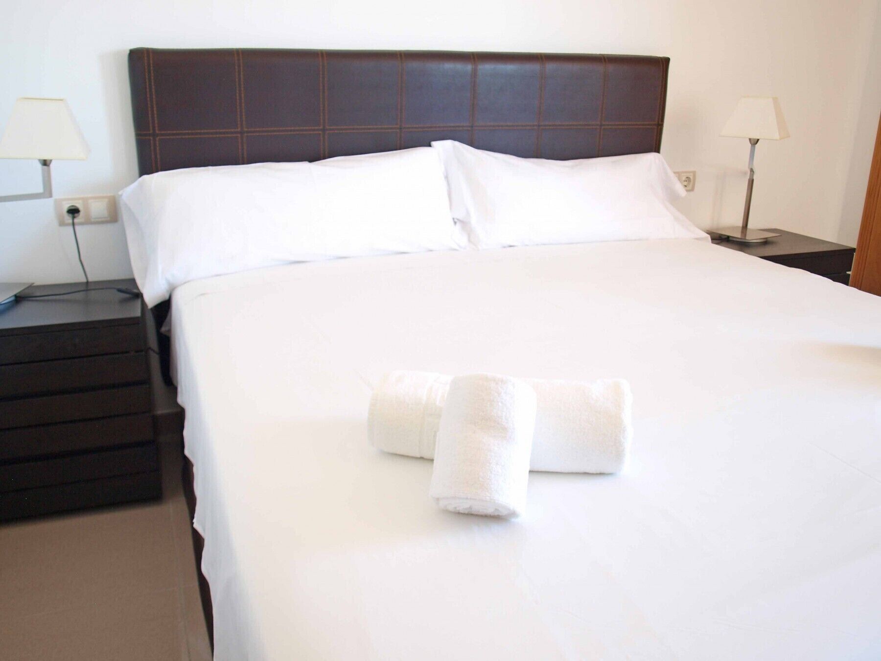 A neatly made bed with white linens and two rolled towels, a dark headboard, and bedside lamps on nightstands.