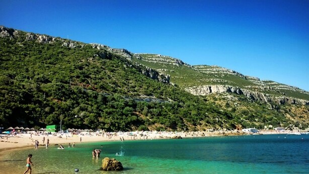 A beach with clear turquoise water, a rocky hillside, and a crowded beach area with umbrellas.