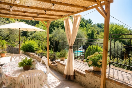Une terrasse extérieure couverte, avec une table et des chaises, entourée de plantes en pot et offrant une vue sur une piscine.. Studio à la campagne avec piscine, vue exceptionnelle, propice à la détente et aux voyages