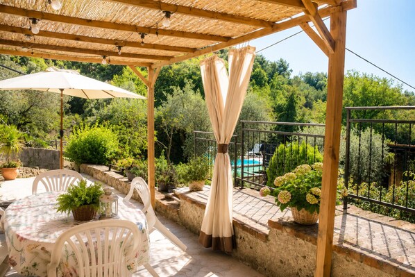 A covered outdoor patio with a table and chairs, surrounded by potted plants and a view of a swimming pool.
