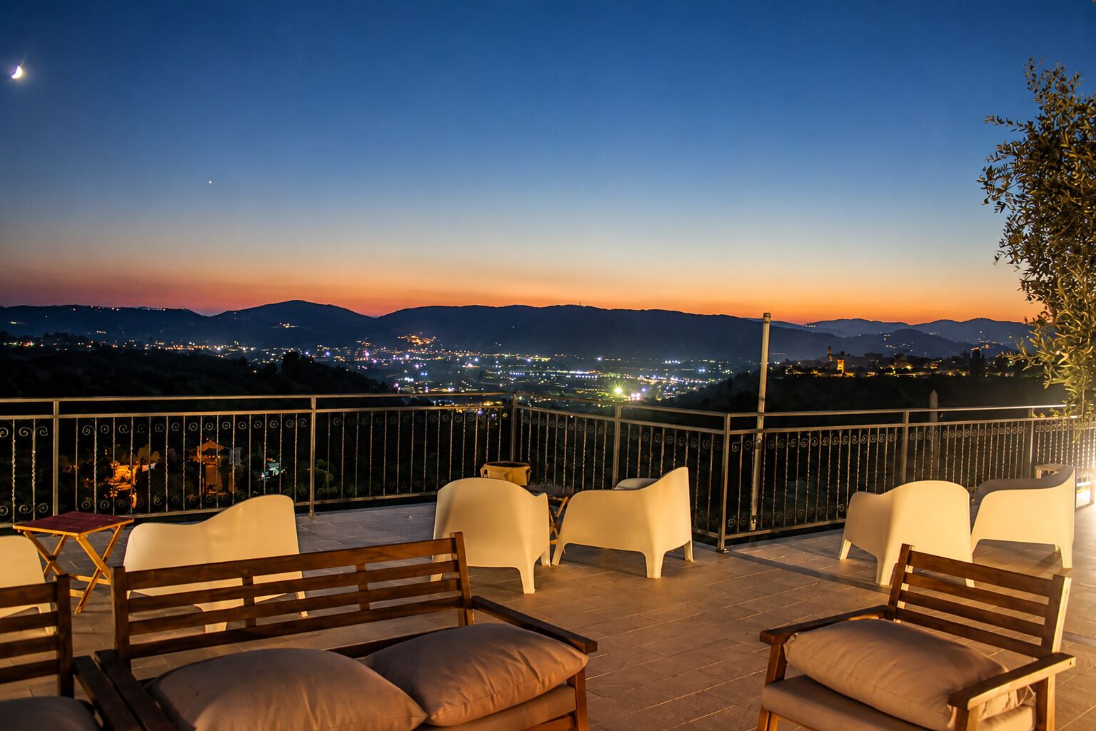 A terrace with outdoor seating, a view of a cityscape at dusk, and a mountain range in the background.