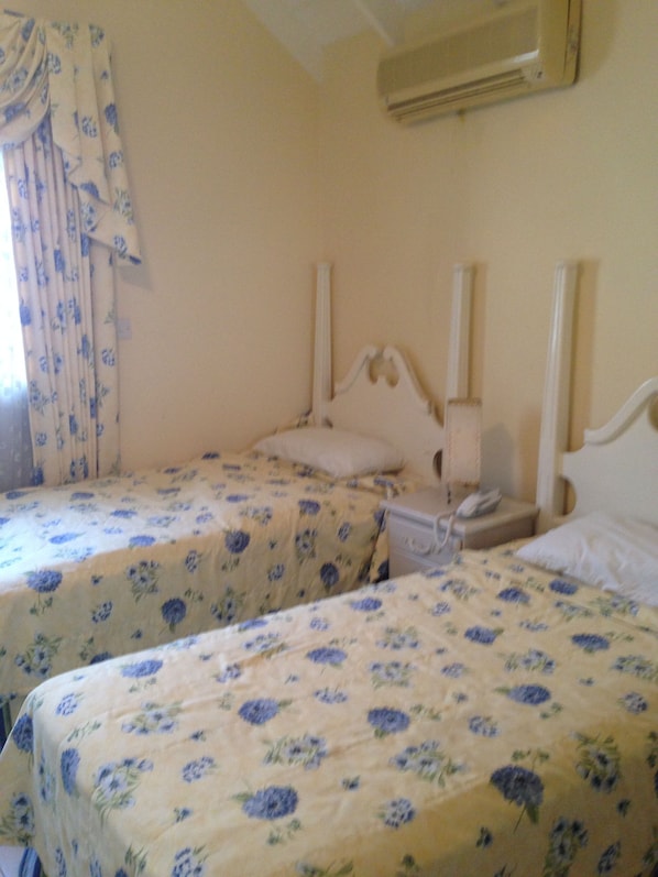 A room with two beds, each with floral bedding, a bedside table with a lamp, and an air conditioning unit on the wall.