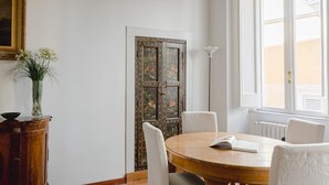 A dining area with a round wooden table, white chairs, a vase with flowers, a framed painting, and a large window with blinds.