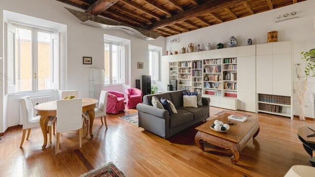 A spacious living room with a wooden ceiling, a grey sofa, a wooden coffee table, and a white bookshelf filled with books.