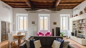 A cozy living room with a dark sofa, white chairs, a wooden dining table, and a bookshelf.