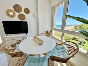A room with a round table, wicker chairs, a flat-screen TV, and large windows with a beach view.