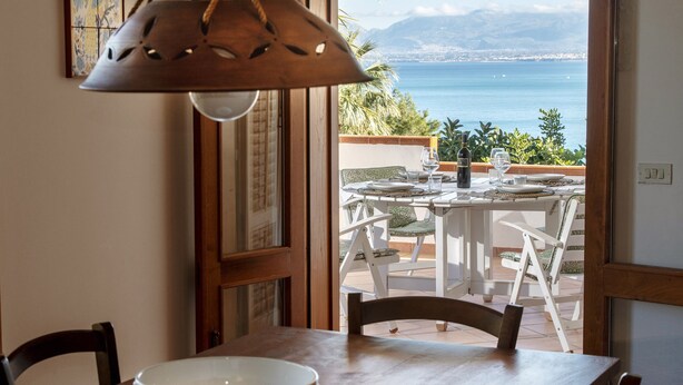 Een eethoek met een tafel gedekt voor twee personen, een hangende lamp en een uitzicht op zee en bergen door een open deur.
