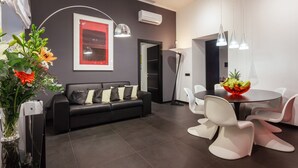 A modern living room with a black sofa, white chairs, a dining table with a fruit bowl, and a vase of flowers. - New duplex apts in VATICAN - M + I (Roma)