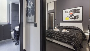 A modern bedroom with a large bed, a bedside table, a wall-mounted artwork, and a bathroom visible through an open door. - New duplex apts in VATICAN - M + I (Roma)
