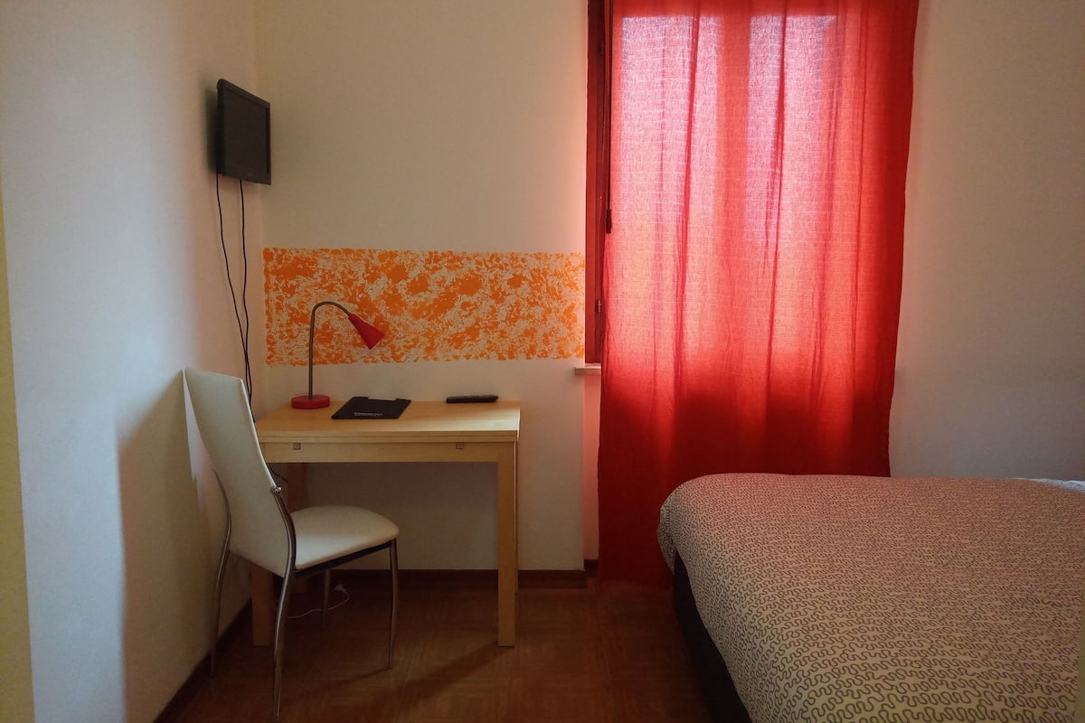 A room with a desk, chair, bed, TV, and red curtains.