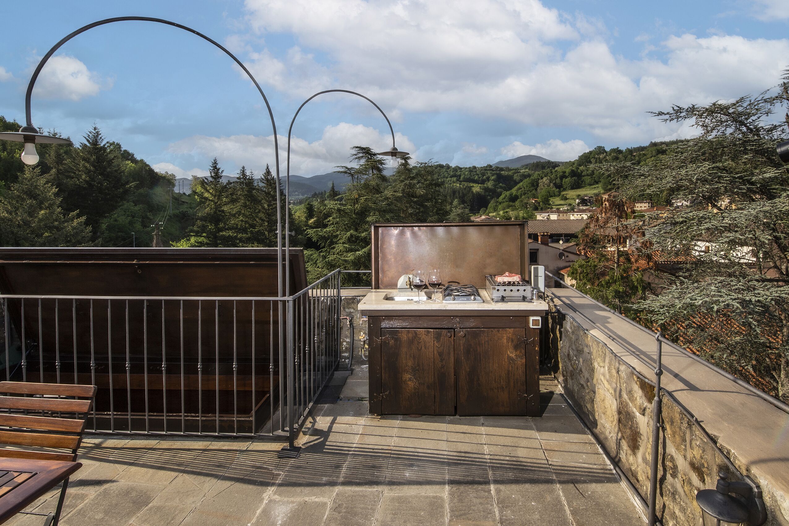 A rooftop terrace with a wooden bench, a small bar area with a sink and wine glasses, and a view of a green hillside with buildings in the distance.