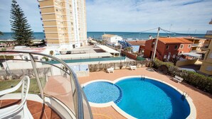 Un espace piscine offrant une vue dégagée sur la mer, un immeuble imposant et une construction au toit rouge.