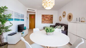 Un salon moderne comprenant une table à manger, des chaises blanches, un téléviseur, une plante, des œuvres d’art et un canapé.