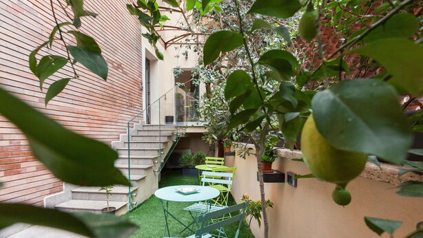 Een kleine binnenplaats met een groen gazon, een witte tafel en twee groene stoelen. Er zijn treden die naar een balkon leiden, en een bakstenen muur aan de zijkant.