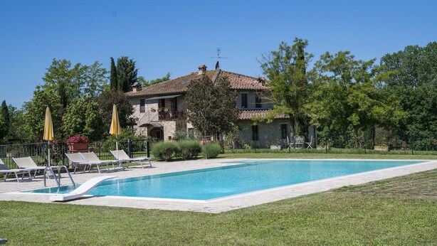 Une maison en pierre à deux étages, dotée d’une piscine, entourée de verdure et baignée d’un ciel dégagé.