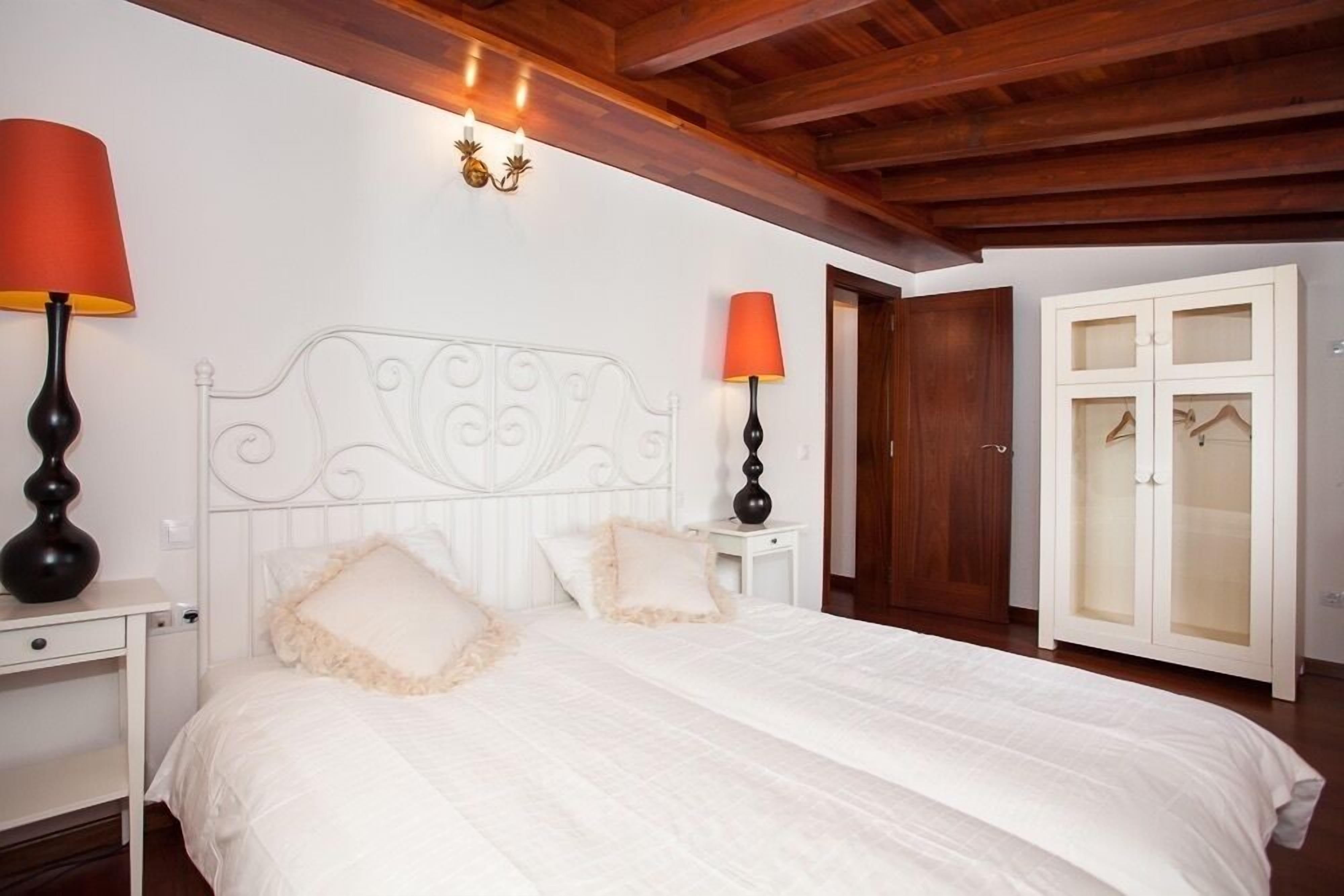 Une chambre à coucher comprenant un lit, deux lampes de chevet, une armoire et un plafond avec des poutres apparentes.