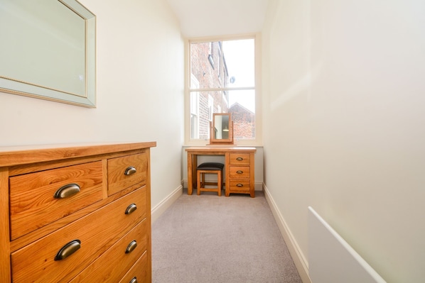 A small, narrow room with a wooden dresser and a desk with a chair. A window provides a view of a brick building outside.