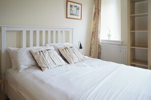 A bed with white bedding and striped pillows, a nightstand, a framed picture on the wall, a window with a plant, and a radiator.