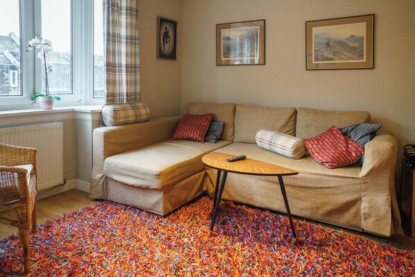 A cozy living room with a sofa, a small table, a colorful rug, and a window with curtains.