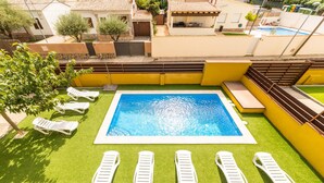Une piscine rectangulaire dont le centre est bleu, entourée de chaises longues blanches et d’une pelouse artificielle verte.