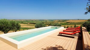 Une piscine rectangulaire sur une terrasse en bois, agrémentée de chaises longues rouges, le tout entouré d’un paysage rural.