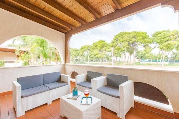 A covered outdoor seating area with a sofa and chairs, a small table with a drink and a book, and a view of trees and greenery. - Apartament 350m from the sea - two verandas - parking - air conditioning (Specchiolla)