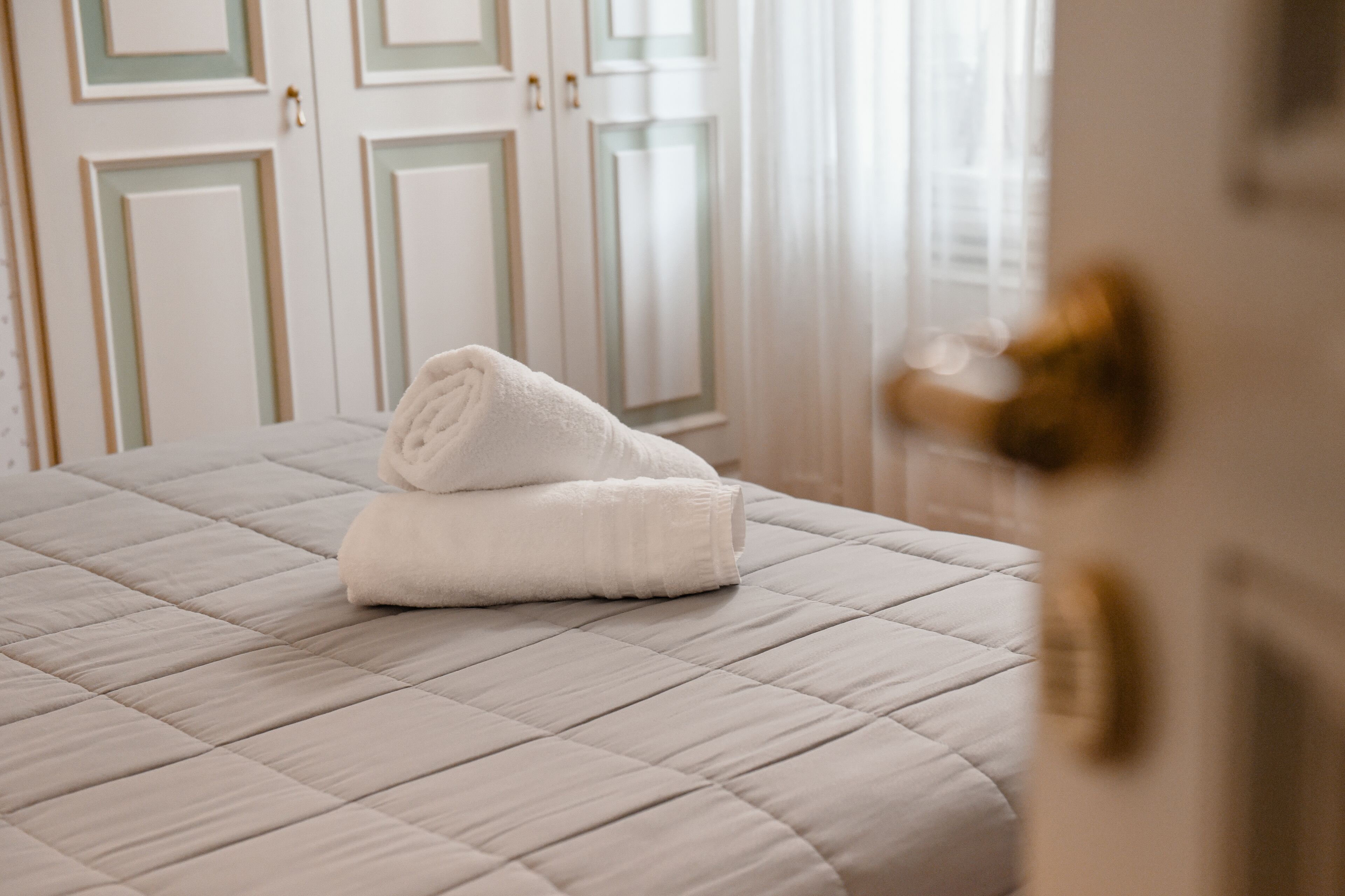A neatly made bed with a folded white towel, a white quilted bedspread, and a door with a brass handle in the foreground.