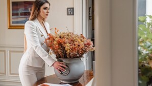 Una mujer con un traje blanco posa junto a un gran jarrón gris con flores secas.