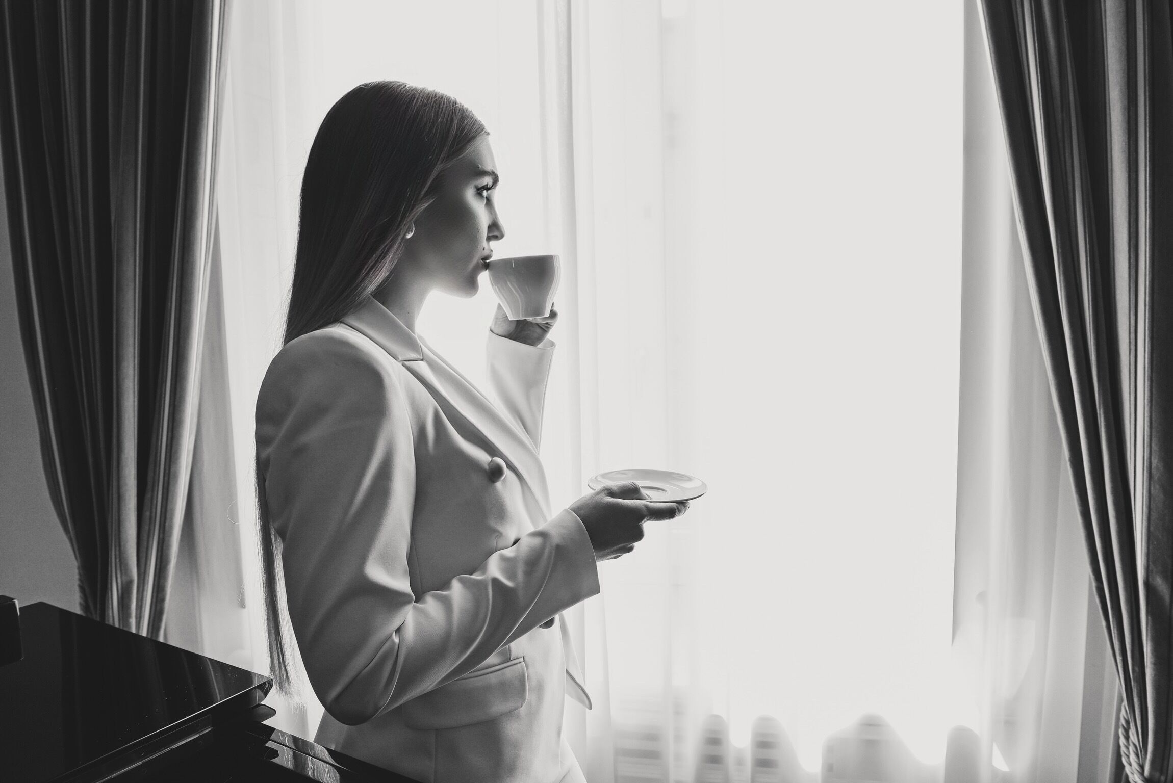 Une personne se trouve dans une pièce où il y a un piano. Elle est debout près d’une fenêtre, derrière laquelle on aperçoit des rideaux, et elle tient une tasse et une soucoupe.