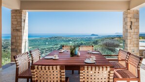 A dining area with a table set for four, surrounded by chairs, overlooking a scenic view of the countryside and sea. - Lux Villa★SPA & Tennis★Jacuzzi & Indoor Pool★BBQ & Gym (Xirokampi)