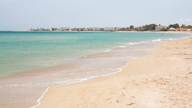 Una playa con aguas turquesas cristalinas, una orilla arenosa y un pueblo lejano con edificios.