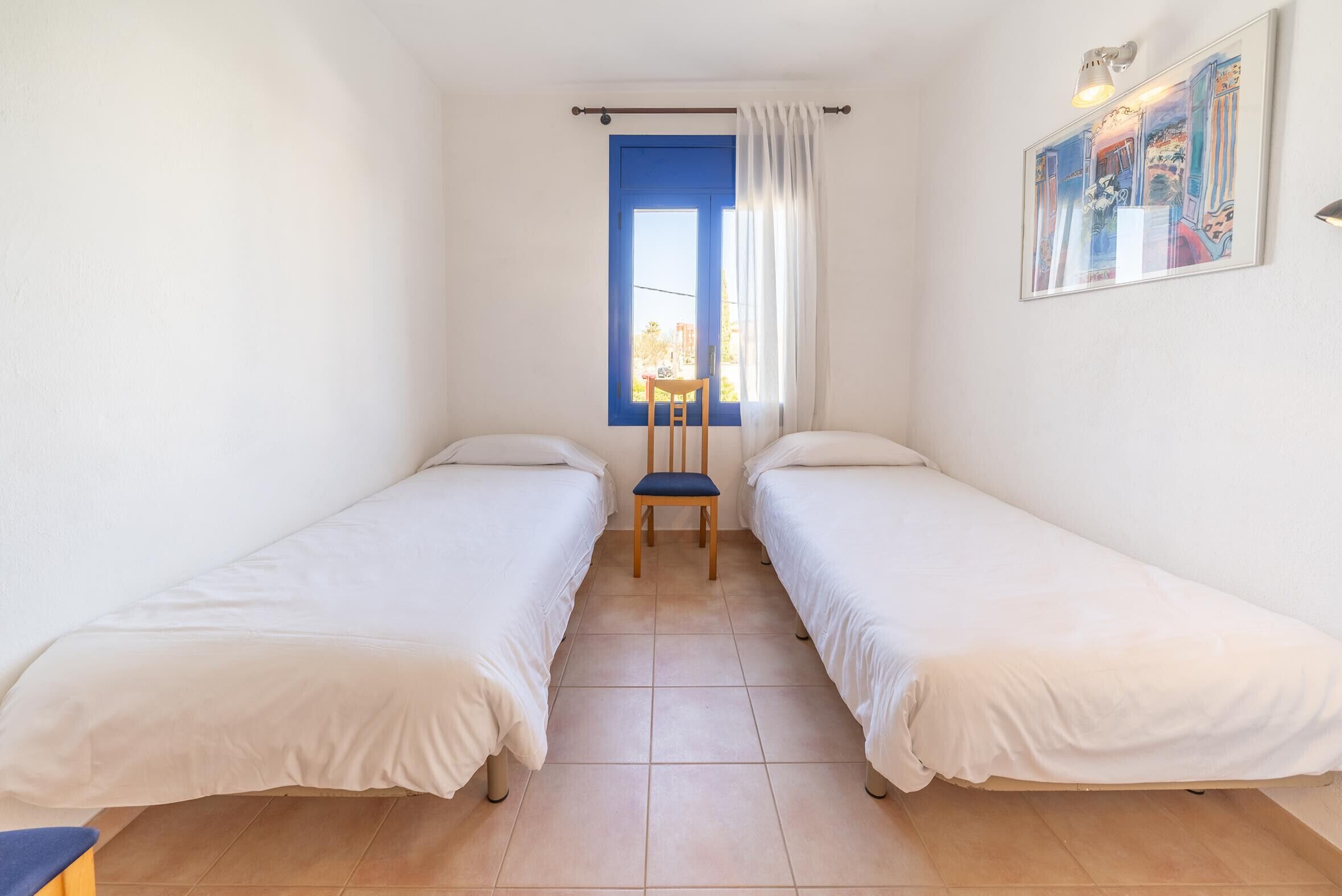 Une petite chambre propre, avec deux lits simples, une chaise, une fenêtre avec des rideaux bleus et un tableau au mur.