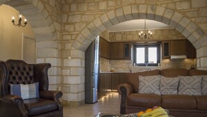 Un vaste salon comprenant un canapé en cuir, un fauteuil à capitons et une table basse en verre sur laquelle sont disposés des fruits. Un arc en pierre mène à une cuisine équipée d’appareils électroménagers modernes et dotée de meubles en bois.