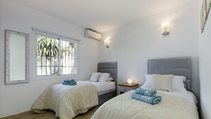 Habitación con dos camas, cada una con cabecero, mesita de noche y lámpara. Tiene una ventana con barrotes, un equipo de aire acondicionado y un espejo en la pared.