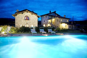 Un edificio in pietra con piscina, sdraio e un'illuminazione esterna calda al crepuscolo.