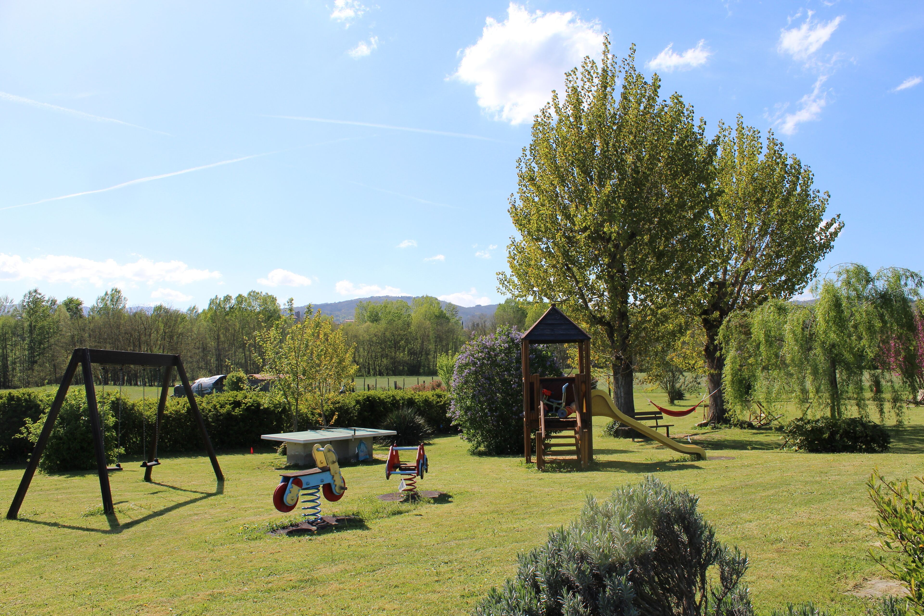 Une aire de jeux avec des balançoires, un toboggan et une structure d’escalade, située dans un espace verdoyant avec des arbres et un ciel dégagé.