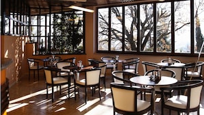 A restaurant with multiple round tables and chairs, large windows with a view of trees, and a tiled floor. - Hotel Eden Spa (Sarnano)