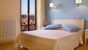 A bedroom with a bed, a nightstand, a lamp, a window with curtains, and a view of a town. - Hotel Eden Spa (Sarnano)