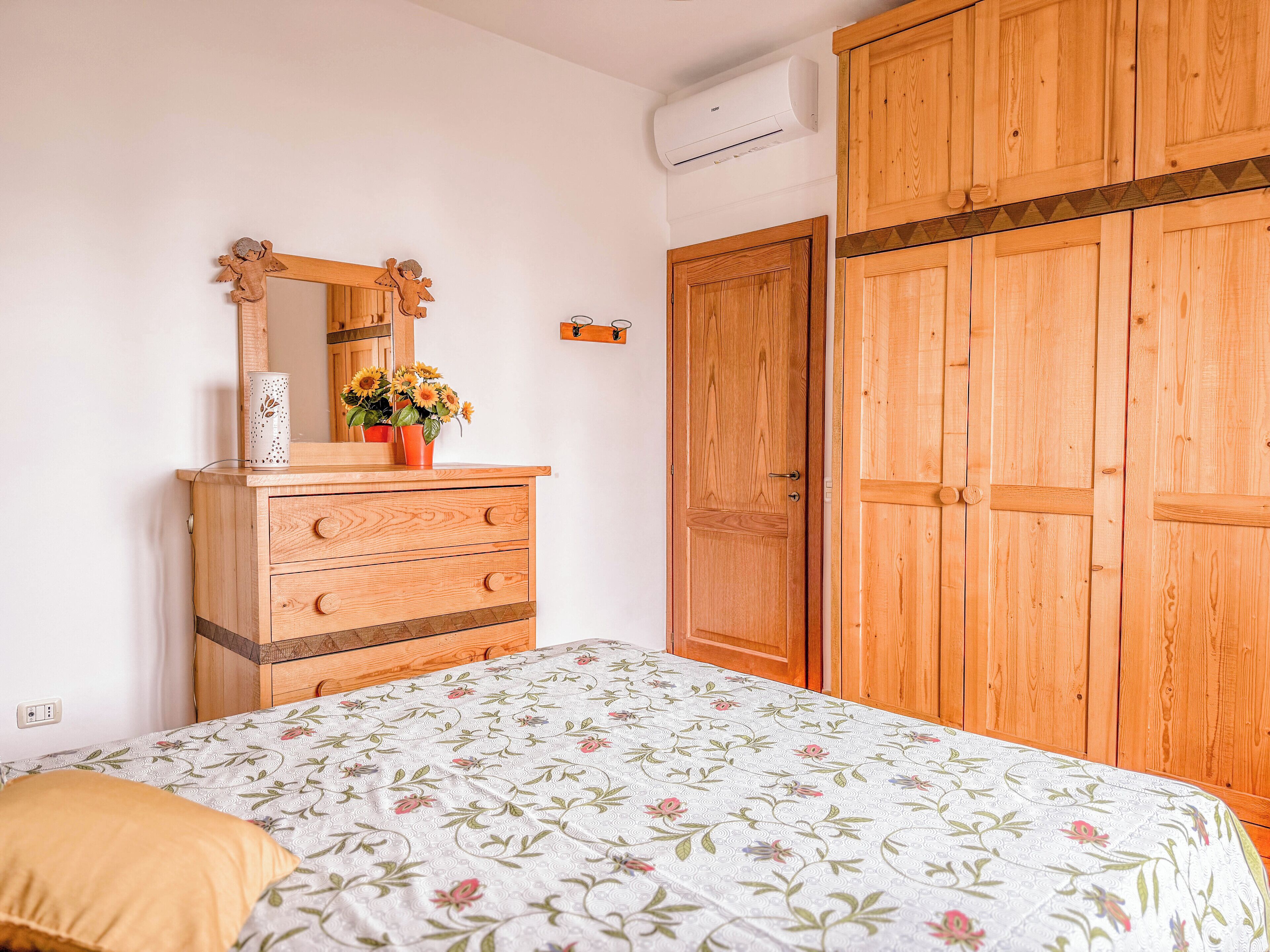 Une chambre à coucher avec une commode en bois, un lit recouvert d’une couvre-lit à motifs floraux, un miroir et une armoire.