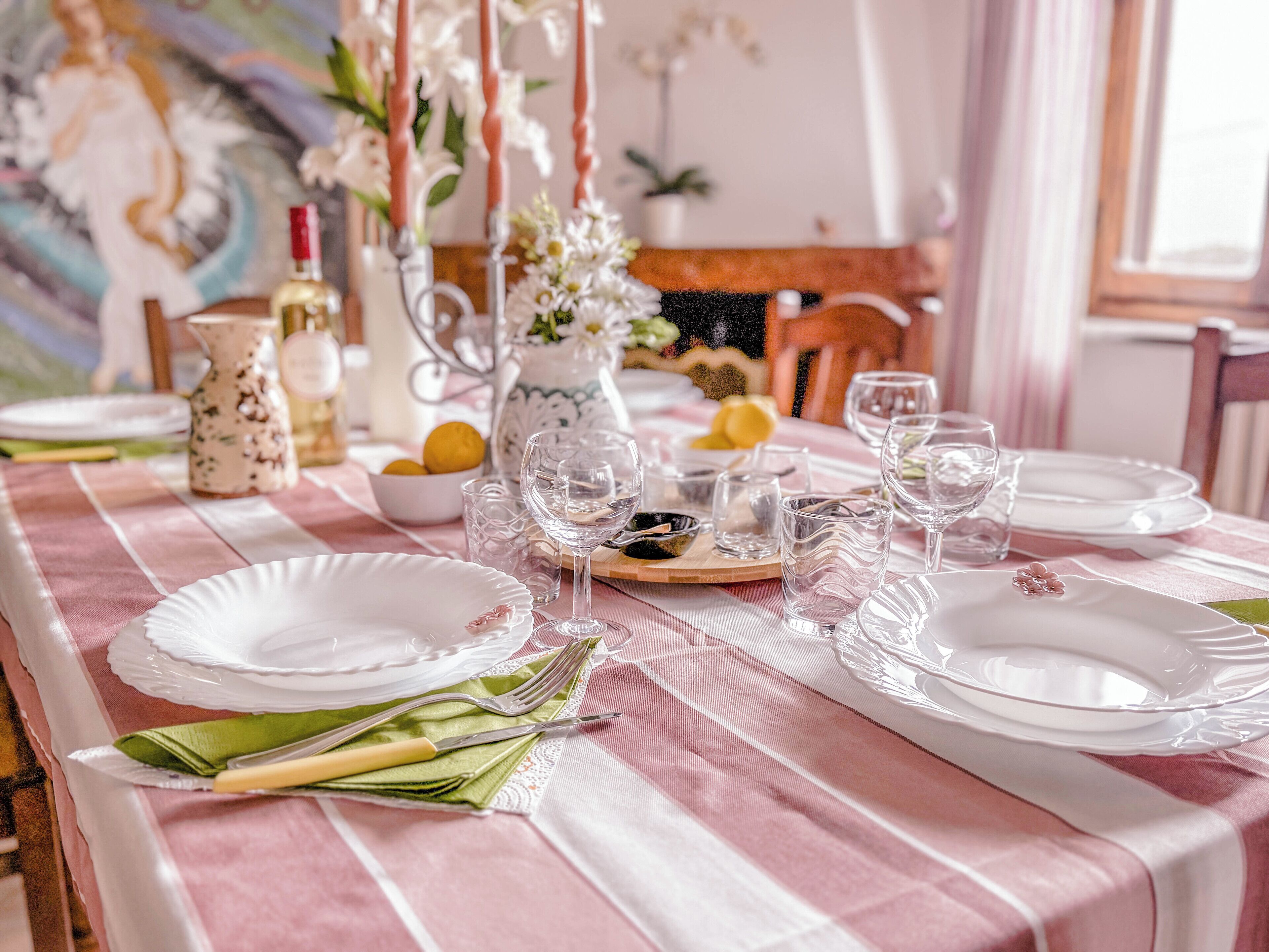 Mesa de jantar posta com pratos, copos e talheres, com um vaso de flores e um jarro de água.