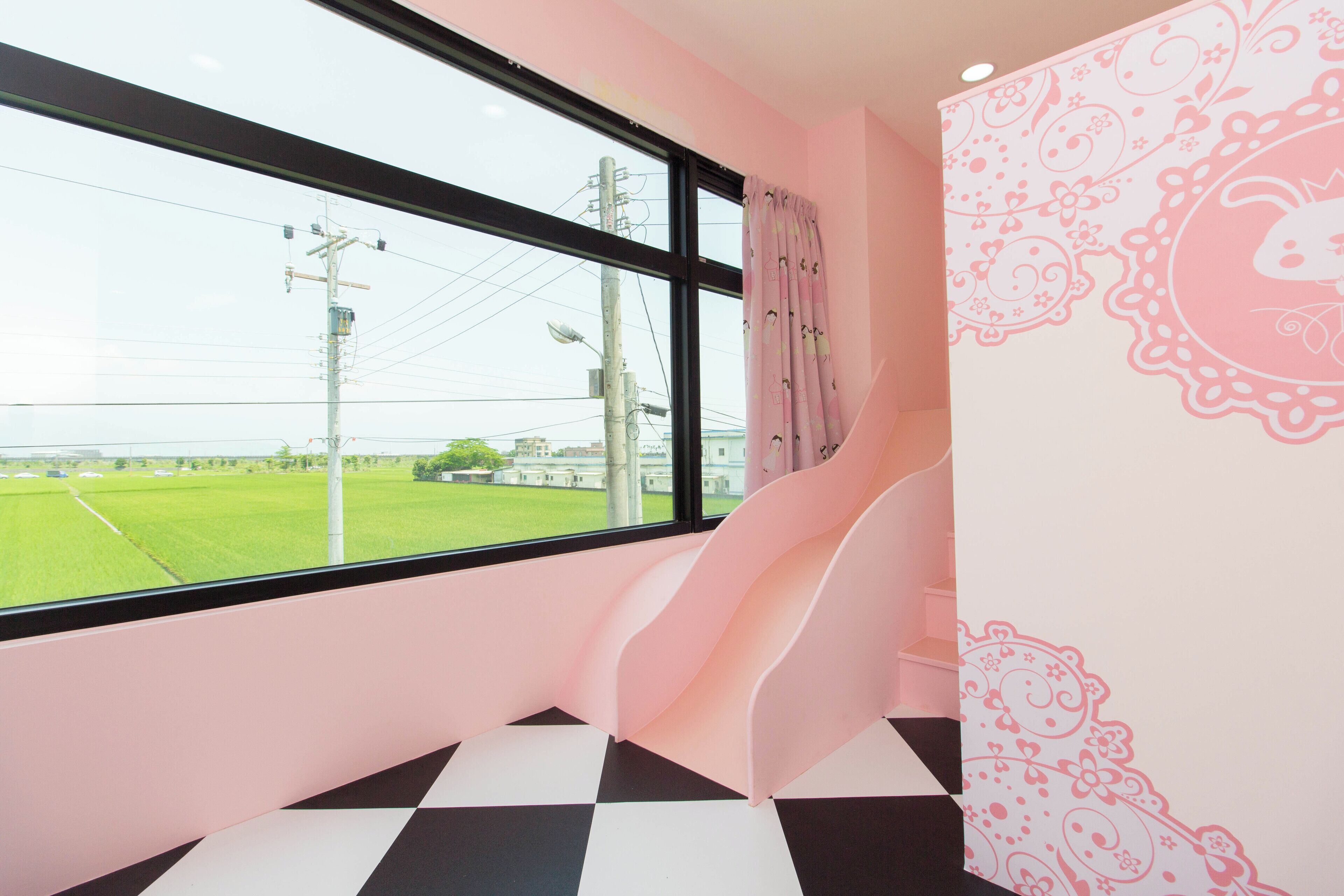 Una habitación con un ventanal grande, una pared rosa con motivos florales y un suelo a cuadros blanco y negro.