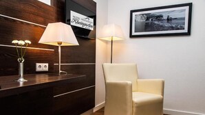 A hotel reception area with a wooden reception desk, two lamps, a framed photograph on the wall, and a cream-colored chair. - Hotel Rheingarten Duisburg (Duisburg)